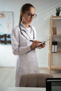 Beyaz önlüklü doktor kadın hastane ofisinde duran hastası için reçete yazıyor. Boşluğu kopyala