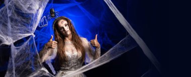 Korkunç, korkunç Cadılar Bayramı zombi makyajlı, beyaz elbiseli, mavi arka planda duran bir şeyi tavsiye eden, örümcek ağıyla çevrili bir kadın. Şeytan insan. Korku filmi. Pankart