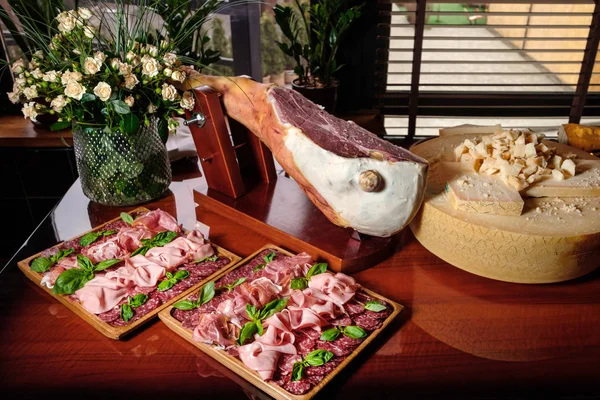 Jamon em uma mesa de madeira com queijo parmesão e carne cortada no fundo de uma janela com jalousie e flores . — Fotografia de Stock