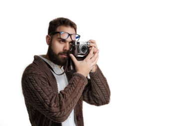 Beyaz arka planda yalnız poz veren kameralı kendine güvenen hippi fotoğrafçı. Koyu kahverengi kazak giyen, siyah sakallı ve gözlüklü bir manken.