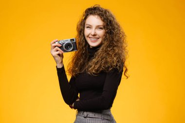 Kıvırcık saçlı, güzel gülüşlü, sarı arka planda izole fotoğraf makinesi olan mutlu bir kız.