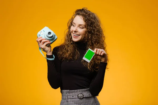 Beautiful girl with instant film camera isolated on yellow background. Model is holding blank frame with mockup space on a picture