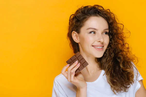 チョコレートバーとかなり笑顔で幸せな女の子 モデルは白い歯を持つ歯列矯正治療のための歯科ブレースを持っています 黄色の背景に隔離された閉鎖的な肖像画白人女性とコピースペース — ストック写真