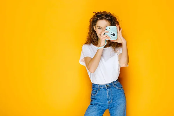 Pretty girl with instant camera isolated on yellow background. Model wearing casual white shirt and has long curly hair.