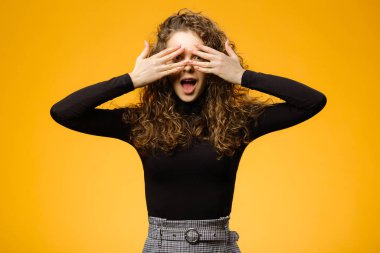 Güzel kız sarı arka planda elleri izole bir şekilde yüzünü gizliyor. Uzun kahverengi kıvırcık saçlı bir model.