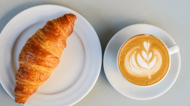 En üstteki tabakta kruvasan ve latte ile beyaz masa üzerinde latte sanatı