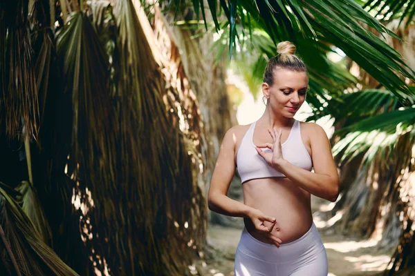 Young attractive blond woman in white sportswear doing yoga pilates fitness exercises in tropical nature. Pre natal exercising, healthy lifestyle, recreation concept. Idyllic scenery for workout