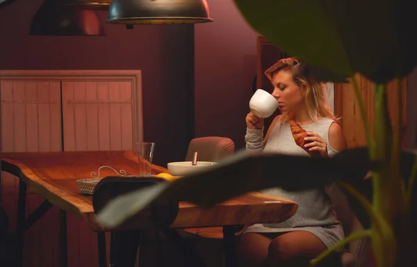 若い金髪の妊婦座っていると 自宅のキッチンでパン屋さんのクロワッサンとコーヒー お茶を飲むだけ — ストック写真