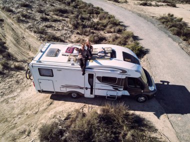 View from above, aerial photo recreational vehicle motor home trailer near empty road. Married couple sitting at car roof and  waving hands sitting at sunbed mattress feels happy having romantic date clipart
