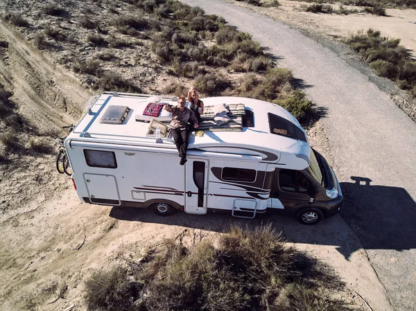 View from above, aerial photo recreational vehicle motor home trailer near empty road. Married couple sitting at car roof and  waving hands sitting at sunbed mattress feels happy having romantic date