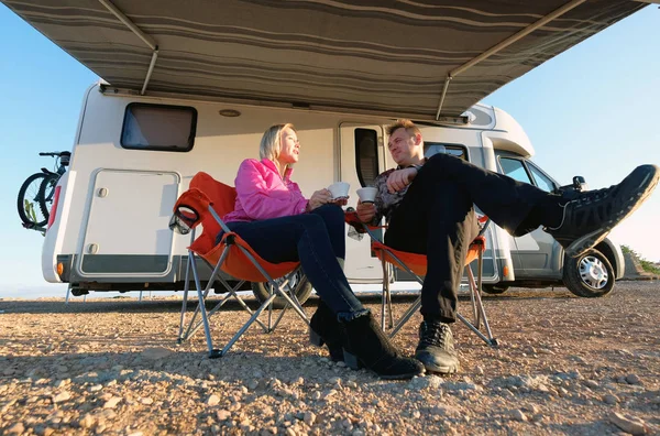 Middle aged married couple wife and husband holding cups drinking tea sitting on folding chairs near recreational vehicle motor home trailer take a break during trip resting talking feel happy