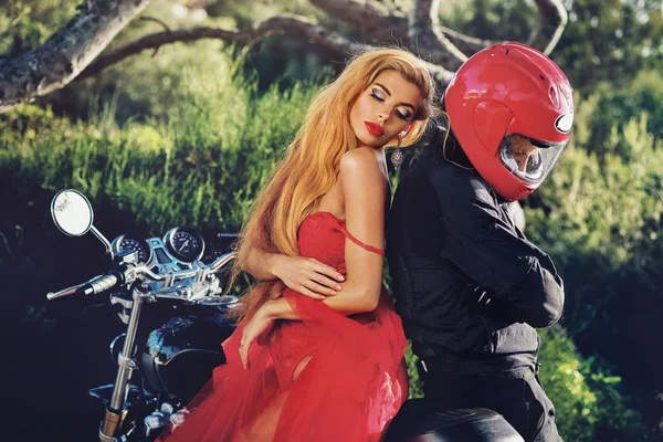 Dame dragen rode jurk zittend op motorfiets met man, meisje dat op Guy verbergt zijn gezicht achter helm pose op de natuur, zonnige zomerdag twee mensen buitenshuis in de buurt van fiets, hobby Lifestyle Fashion concept — Stockfoto