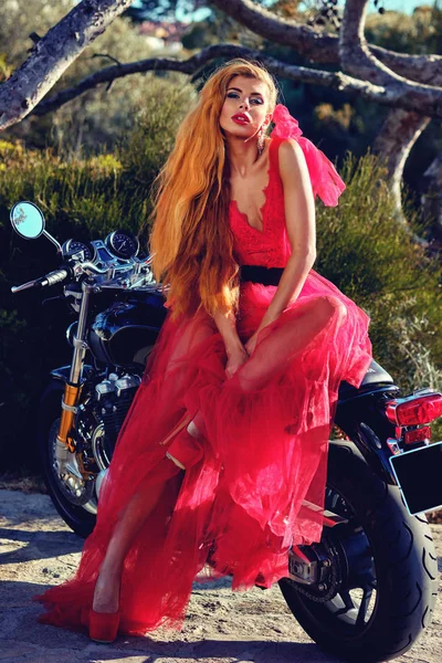 Vacker kvinna i röd fluffiga klänning poserar sittande på motorcykel — Stockfoto