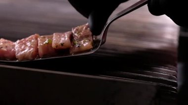 Şef balık dilimleri pişiriyor, siyah eldivenli adam metalik spatula tutuyor terbiye edilmiş cerrahların parçalarını barbekü ızgarasının sıcak yüzeyine koyuyor, kamerayı yavaş çekimde çekiyor.