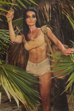 Orta yaşlı sporcu kadınlar seksi şortlar giyer. Sırt çantasında palayla tropik palmiye ağaçlarında poz verirler. Macera ruhu. Hayatta kalan kadın konsepti