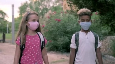 Çoklu etnik arkadaşlar, okul çağındaki sevimli çocuklar okula yürürken konuşurlar. Sırt çantalı ve yüz maskeli çocuklar ders çıkışı dışarı çıkıyorlar. Arkadaşlık ve eğitim kavramı
