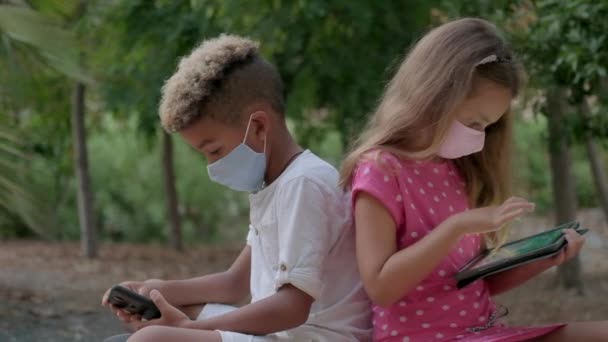 两个9岁10岁的多种族孩子背着面具坐在长椅上 背靠背地使用手机 平板电脑互相忽略 Phubbing 新一代人沉迷于现代技术概念 — 图库视频影像