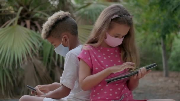 两个9岁10岁的多种族孩子背着面具坐在长椅上 背靠背地使用手机 平板电脑互相忽略 Phubbing 新一代人沉迷于现代技术概念 — 图库视频影像