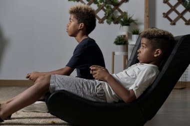 Two mixed race brothers play to play station seated together indoor. Alpha generation offspring addicted with virtual games problem concept clipart