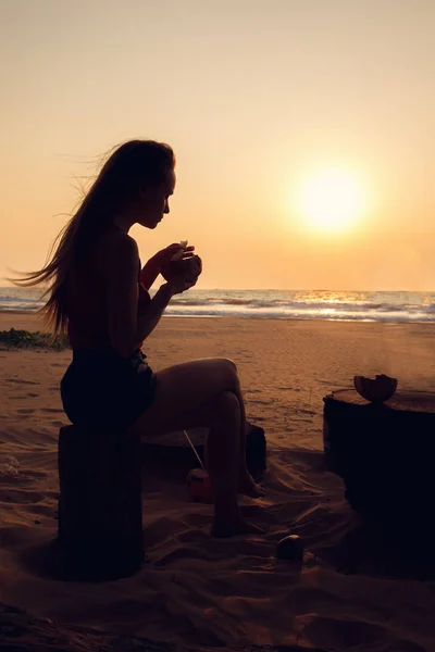 Genç Kadın Hindistan Cevizi Okyanus Kıyısında Günbatımı Yiyor — Stok fotoğraf