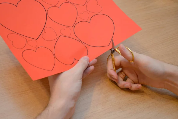 Close-up of a man cutting hearts out of red paper according to a pattern with small golden scissors