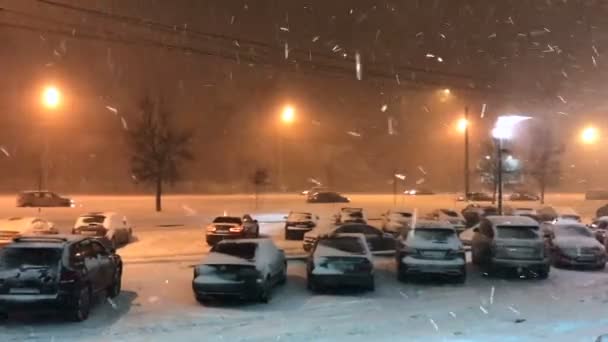 在傍晚的城市里 在灯笼的灯光下 汽车在大雪下沿着公路缓慢行驶 在前景中 被雪覆盖的汽车停在停车场 侧视图 — 图库视频影像