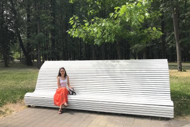 Beyaz gömlek li ve uzun turuncu etekli beyaz genç esmer kadın parkta beyaz bir bankta oturuyor ve güneşli bir yaz gününde kameraya bakıyor..