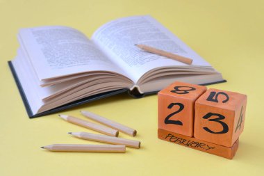 Sürekli ahşap takvim tarihi 23 Şubat, açık bir kitap ve kalemler üzerinde kopya alanı ile sarı bir arka plan ile. Defender Anavatan gün, cesaret, güç kavramı. Yatay fotoğraf