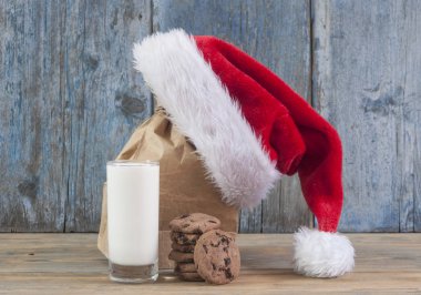 Bardak süt, lezzetli çikolatalı kurabiye ve ahşap masa arka plan üzerinde Santa Claus şapka