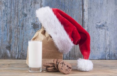 Bardak süt, lezzetli çikolatalı kurabiye ve ahşap masa arka plan üzerinde Santa Claus şapka