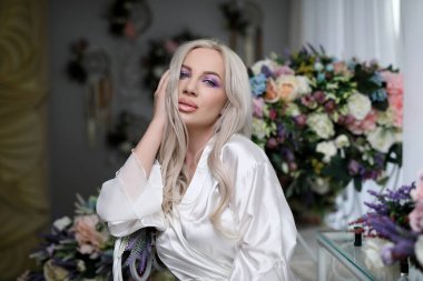 Yatak odasındaki kız. Beyaz ipek bornozlu güzel bir sarışın. Çiçeklerin arka planında, kameraya bakıyor..