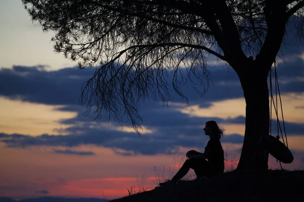 快乐的年轻女子坐在树下欣赏日落的剪影 — 图库照片