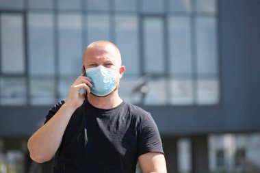 COVID-19 Pandemik Coronavirüs. İş adamı telefonu modern binanın önünde kullanıyor. SARS-CoV-2 virüsünün yayılması için maske takıyor.. 