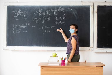 Maskeli bayan öğretmen okulda matematik dersi veriyor. Coronavirus salgını sırasında sosyal uzaklık ve sınıf güvenliği