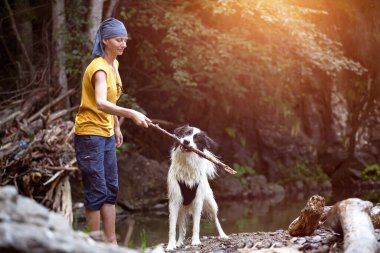 Genç bir kadın ormandaki sığ nehir suyunda köpeğiyle oynuyor..