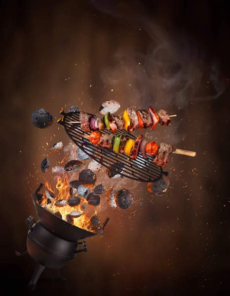 Parrilla para hervidor con briquetas calientes, rejilla de hierro fundido y sabrosos pinchos volando en el aire . — Foto de Stock