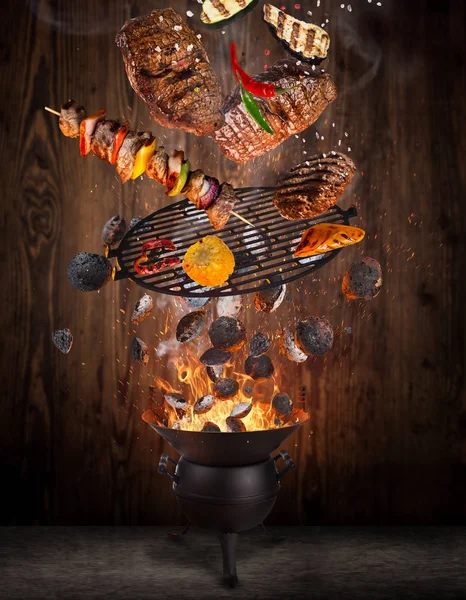 Grelha de chaleira com briquetes quentes, grelha de ferro fundido e bifes saborosos voando no ar . — Fotografia de Stock
