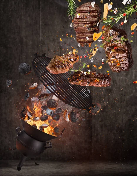 Kettle grill with hot briquettes, cast iron grate and tasty beef steaks flying in the air.