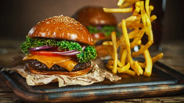 Frytki spadają obok cheeseburgera, leżącego na drewnianej desce do krojenia vintage. — Zdjęcie stockowe