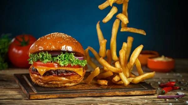 Batatas fritas caem ao lado de cheeseburger, deitado na placa de corte de madeira vintage. — Fotografia de Stock