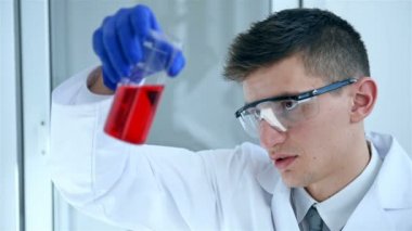 Mavi eldiven ve önlük içinde genç bilim adamı incelemek Flask kırmızı sıvı onun kol ağır çekim etkisi ile bakıyor.