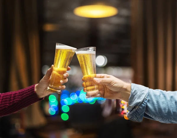 Beer glasses raised in a toast. Close-up hands with glasses. Blurred bar interior at the background.