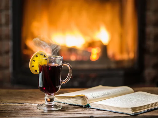 ホットホット ワインと木製テーブルの上の本 背景に暖かい火が付いている暖炉 — ストック写真