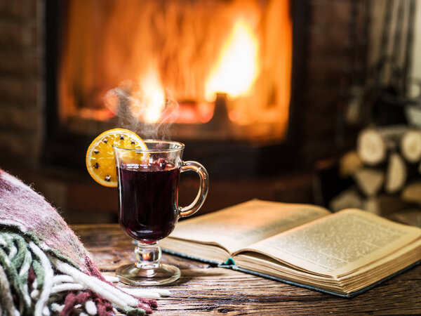 Hot mulled wine and a book on the wooden table. Fireplace with warm fire on the background.