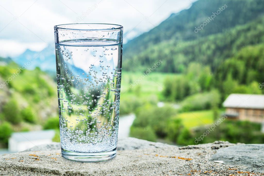 Glass of water on the stone. Blurred snow mountains tops and green forests at the background, as a symbol of freshness and purity.