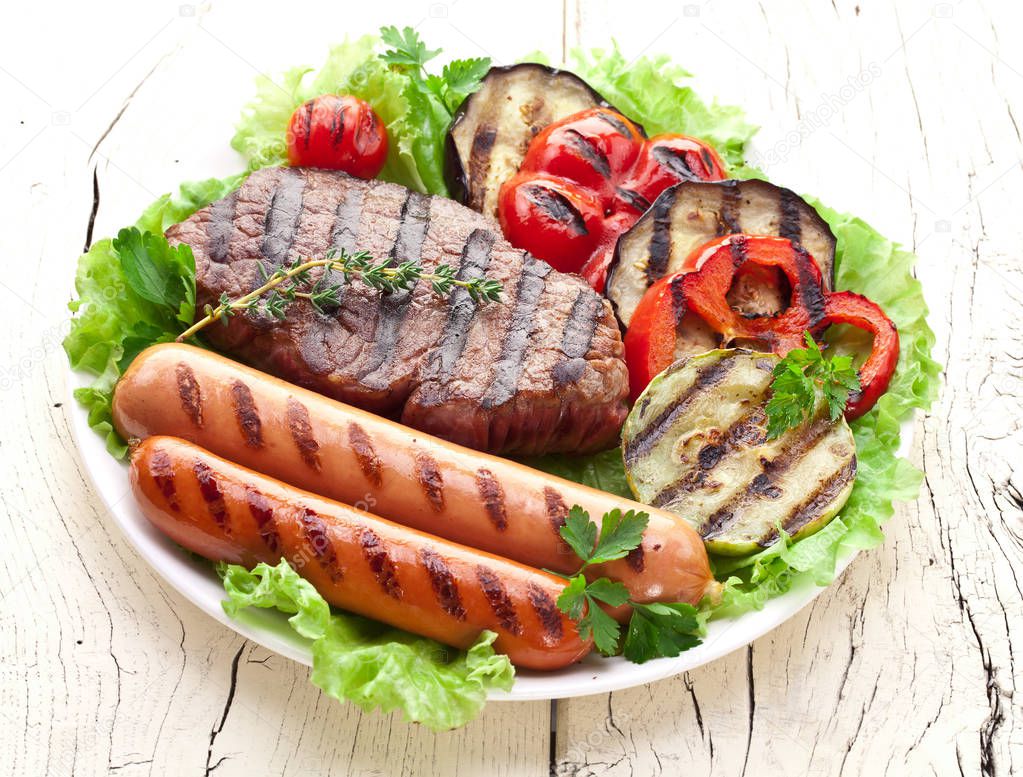 Grilled meat and vegetables over green leaves on white plate on the wooden table.