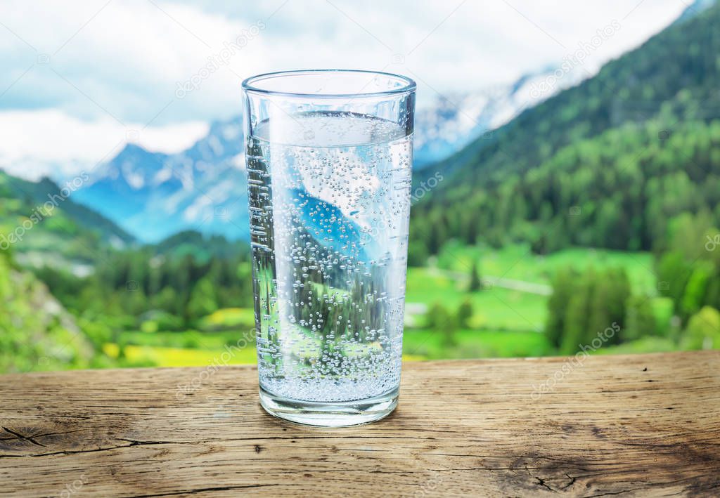 Glass of water on the stone. Blurred snow mountains tops and green forests at the background, as a symbol of freshness and purity.