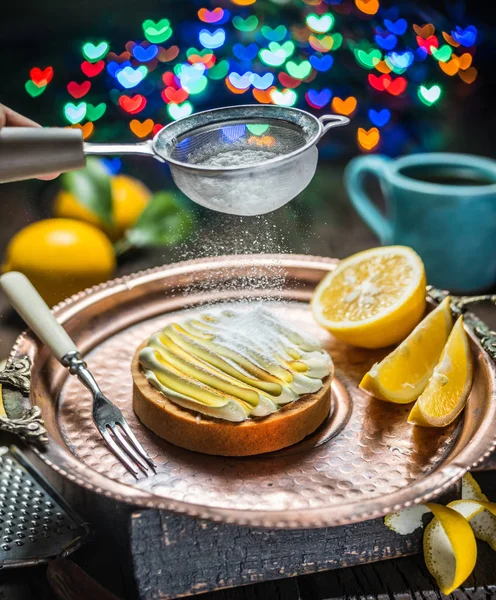 Lemon tart. Cooking of lemon dessert. Sugar powder is sprinkled on top through a strainer. Blurred heart festive background.