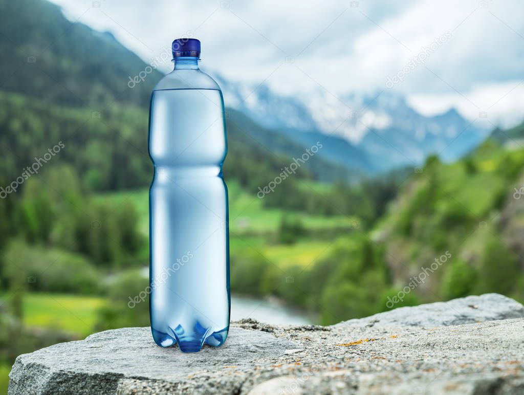 Bottle of water on the stone. Blurred snow mountains tops and green forests at the background, as a symbol of freshness and purity.