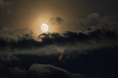 Gece gökyüzünde ağır bulutlar arasında sarı ay.
